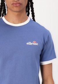 Modré bavlněné tričko s bílým žebrovaným límcem a manžetami na rukávech, s malým vyšívaným logem na prsou.