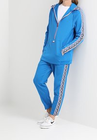 Modrý sportovní teplákový komplet se zipovou kapucí a kalhotami ve stejném stylu. Má přední kapsu a vzorované proužky po stranách. Doplněný bílými teniskami.