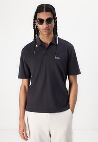Črna polo majica z belim črtastim ovratnikom, kratkimi rokavi in majhnim logotipom "Hollister" na prsih. Model nosi temne sončne očala.