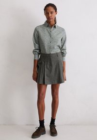 Koszula w kwiatowy wzór w odcieniach zieleni, z zapięciem na guziki i długimi rękawami, zestawiona z plisowaną szarą spódnicą i ciemnymi butami.