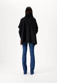 Poncho noir à côtes avec un col montant, présentant une coupe ample et un ourlet asymétrique, associé à un jean évasé bleu. Texture lisse.