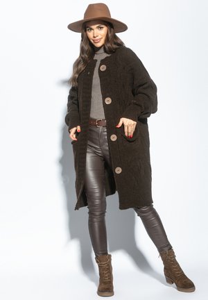 Brązowy sweter z dzianiny z wzorem w warkocze, z dużymi guzikami, zestawiony z szarym golfem, brązowymi skórzanymi spodniami oraz brązowymi botkami na sznurowadła.