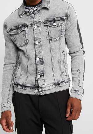 Homme portant une veste en jean gris clair délavée avec des boutons et des rayures noires sur les manches, associée à un pantalon noir.