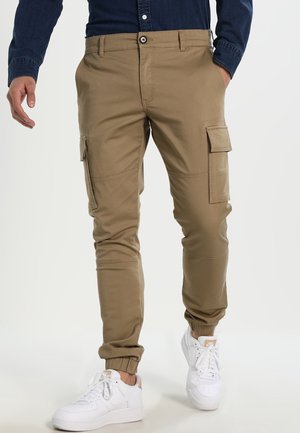 Homme portant un pantalon cargo kaki et des baskets blanches, avec les mains partiellement visibles et une chemise bleu foncé rentrée.