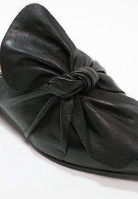 Chaussure plate en cuir noir avec un grand nœud torsadé sur le devant et un bout arrondi lisse.