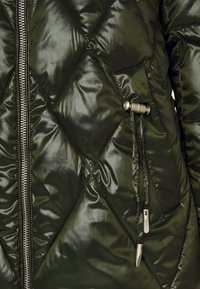 Gequilte olijfgroene pufferjas met een glanzende afwerking, zilveren rits en elastische trekkoord met metalen hardwaredetails.