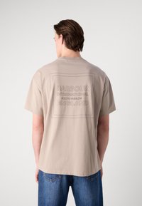 Tricou bej cu mânecă scurtă, prezentând un design de logo gri dreptunghiular pe spate, cu textul "Barbour International South Shields England".