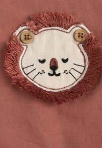 Patch en broderie de lion sur un tissu doux de couleur rose ; présente une crinière à franges, un œil en bouton et des détails faciaux brodés. Design circulaire.