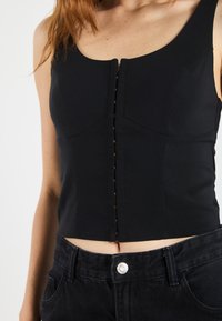 Svart tettsittende topp med scoop-hals, med en frontlukking med knapper. Laget av et glatt stoff, har den en cropped lengde og sømdetaljer.