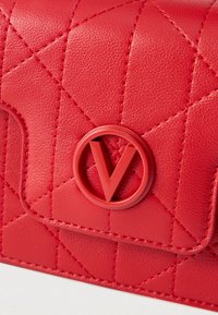 Crveni kožni neseser s uzorkom šavova i utisnutim okruglim logotipom na prednjoj strani. Ima teksturiranu površinu i šavove u istoj boji.