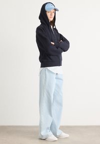 Sudadera navy con cremallera y una capucha grande, gorra azul claro, camisa blanca y pantalones de mezclilla anchos azul claros, combinados con zapatillas blancas.