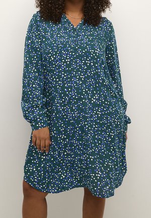 Woman wearing a long-sleeve, green dress with blue, white, and purple dotted pattern and a gold chain necklace.