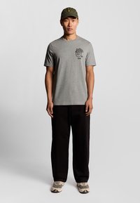 Grijze t-shirt met korte mouwen en een zwarte graphic, gecombineerd met zwarte wijde broek en beige sneakers; groene pet met embleem aan de voorkant.