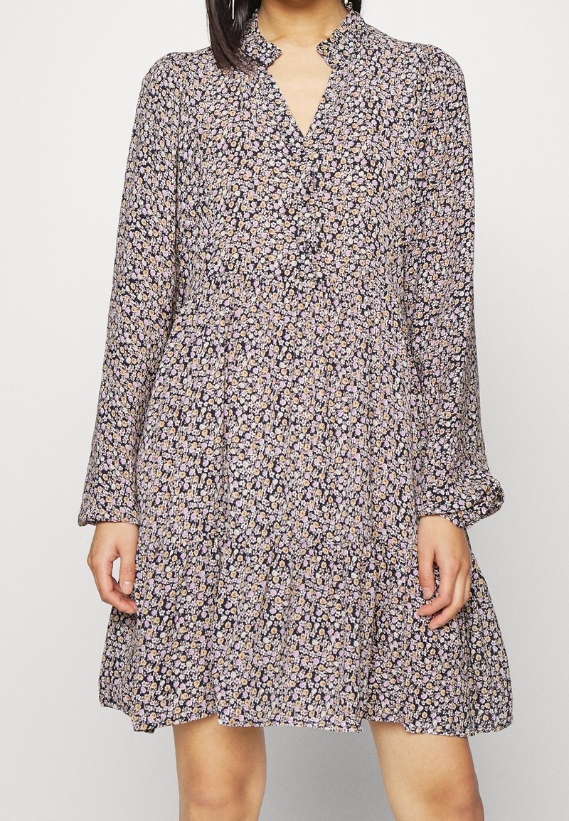 Femme portant une robe noire à manches longues, longueur genou, avec de petits motifs floraux roses et blancs et un col à volants.