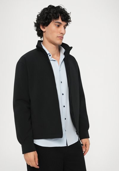Čierna zipsová bunda, mäkká tkanina, voľný strih, vysoký golier, spárovaná s svetlomodrou košeľou s gombíkovou légou a manžetami.
