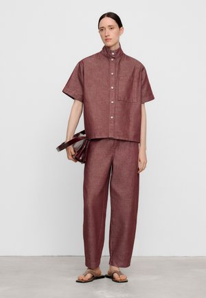Femme portant une chemise ample bordeaux à manches courtes avec boutons et un pantalon assorti, tenant un sac bordeaux, debout sur un fond uni.