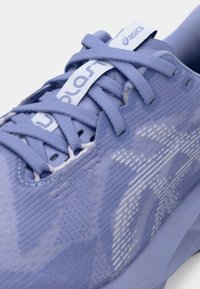 Lyselilla atletisk sko med et teksturert stoffoverdel, fremtredende snøring og en glatt gummisåle med subtile designmønstre.