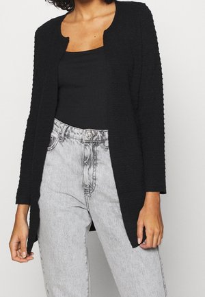 Femme portant un cardigan noir texturé ouvert sur le devant, par-dessus un débardeur noir côtelé et un jean taille haute gris clair.