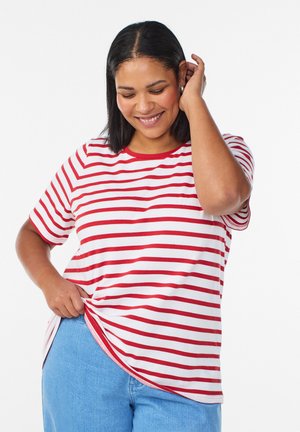 Lächelnde Frau mit mittlerer Hautfarbe und schwarzen Haaren trägt ein rot-weiß gestreiftes T-Shirt und blaue Jeans, während sie ihr Hemd mit einer Hand richtet.