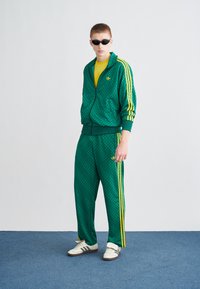 Πράσινο αθλητικό σετ με σχέδιο και κίτρινες ρίγες, που περιλαμβάνει ζακέτα με φερμουάρ και ταιριαστό παντελόνι. Φοριέται με κίτρινη μπλούζα και γυαλιά ηλίου.