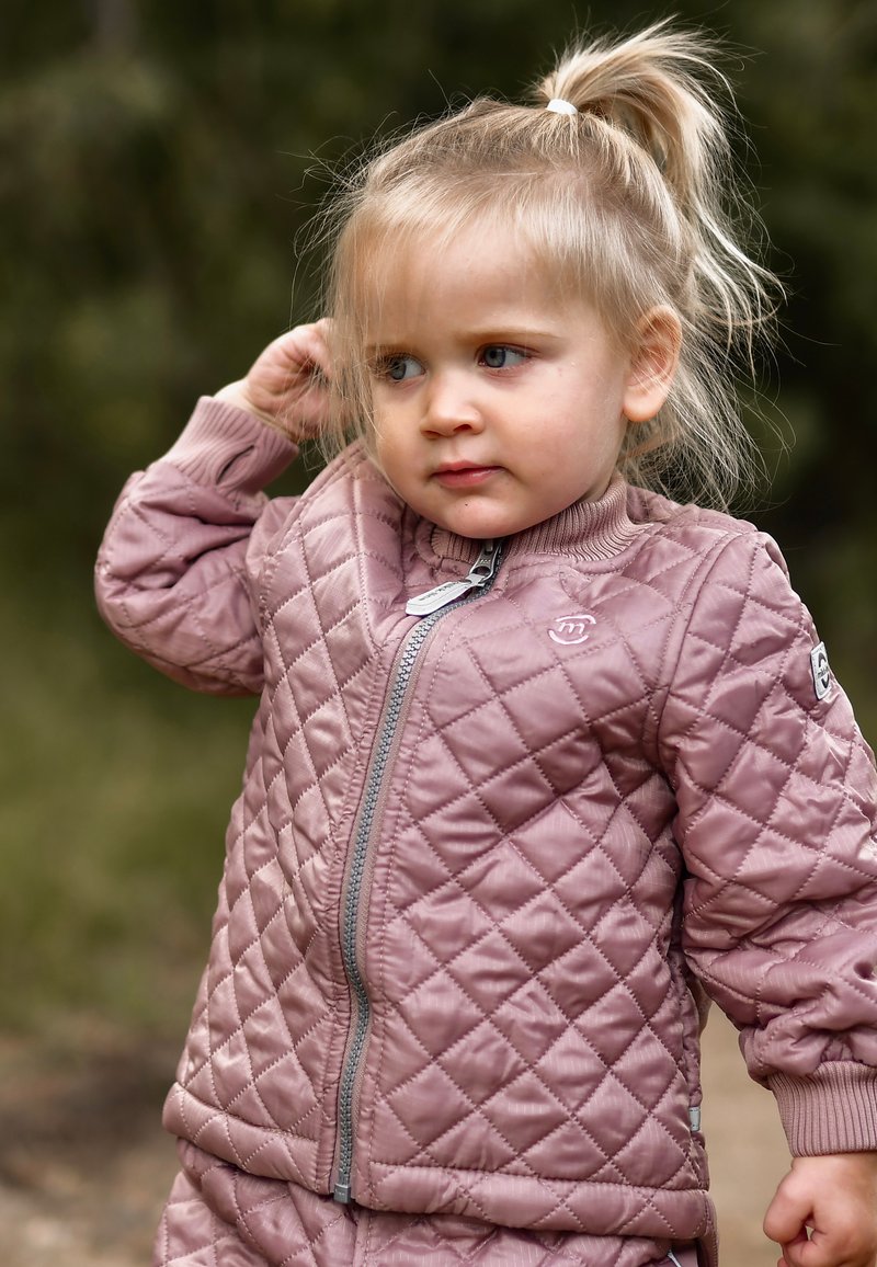 Vatteret lyserød jakke med lynlåslukning, ribbet krave og tekstureret diamantmønster. Modellen har en hestehale og er udendørs.