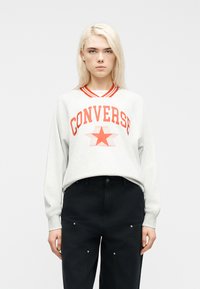 Νεαρός με κανονικά ξανθά μαλλιά, φορώντας λευκό φούτερ με κόκκινο κείμενο "CONVERSE" και αστέρι, συνδυασμένο με μαύρο παντελόνι.