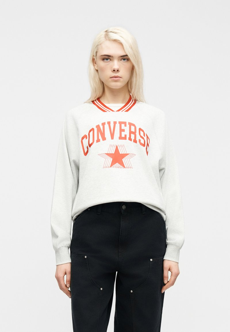 Νεαρός με κανονικά ξανθά μαλλιά, φορώντας λευκό φούτερ με κόκκινο κείμενο "CONVERSE" και αστέρι, συνδυασμένο με μαύρο παντελόνι.