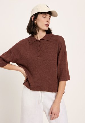 Frau trägt ein braunes, kurzärmeliges, gestricktes Poloshirt, weiße Hose mit Kordelzug und eine beige Baseballkappe, posiert mit einer Hand in der Hüfte.