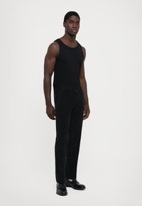 Joven de pie, vistiendo una camiseta sin mangas negra, pantalones negros y zapatos de cuero negros contra un fondo blanco liso.