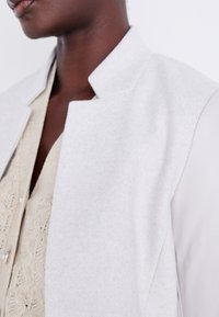 Giacca in misto lana grigia chiara con colletto strutturato e tessuto liscio, indossata sopra un top in maglia beige con un motivo dettagliato.