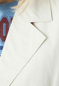 Tela de gabardina beige claro con un solapa prominente y costuras limpias, superpuesta sobre una camiseta gráfica azul con texto rojo.