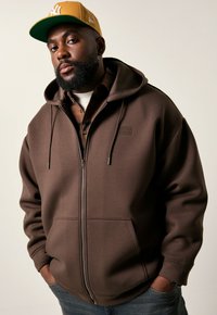 Mann mit Bart, der eine braune Zip-Hoodie, ein kariertes Hemd darunter, eine gelbe Baseballmütze und blaue Jeans trägt, steht mit den Händen in den Taschen.