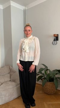 Blusa branca com folhos, mangas compridas e fecho frontal de botões, combinada com calças pretas largas. Tecido macio e design casual. Ambiente interior.