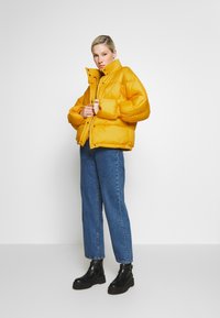Giacca a vento gialla con colletto alto, caratterizzata da una texture lucida e bottoni a pressione, abbinata a jeans in denim blu e stivali neri.