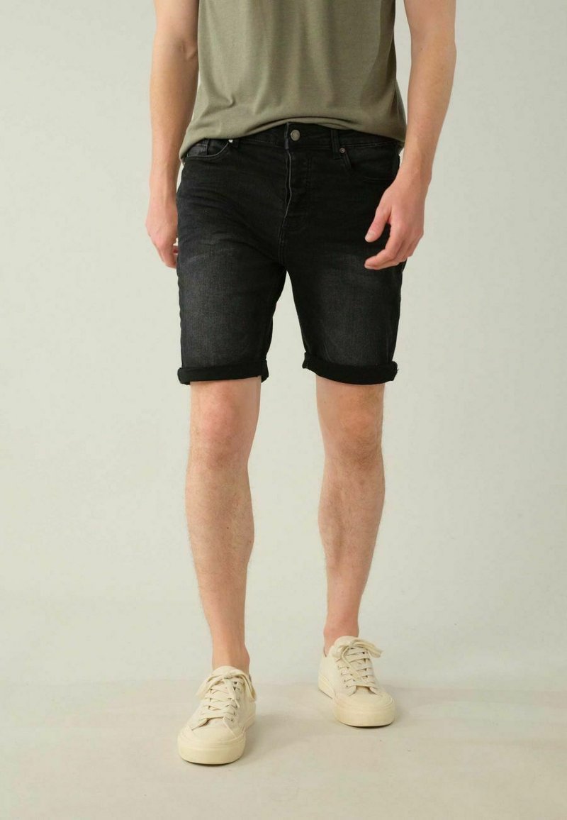 Shorts en denim noir avec ourlets retroussés, coupe slim, associés à un t-shirt ajusté vert olive et des baskets de couleur claire.