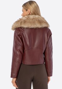 Veste en cuir bordeaux raccourcie avec un large col en fausse fourrure, présentant des manches structurées et une texture lisse, portée sur un pantalon marron foncé.