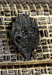 Accessorio nero testurizzato con un dettagliato design della testa di un animale e accenti in pietra nera, su uno sfondo a motivo dorato e nero.