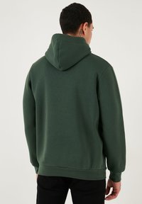 Zelená mikina s kapucňou z textúrovaného materiálu s klokaniajm vreckom, rebrovanými manžetami a voľným strihom. Pohľad zozadu neukazuje žiadne výrazné vzory.