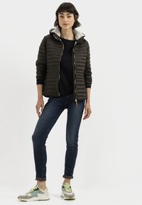 Gevoerde donkerbruine jas met een lichtgrijze capuchon, een zwarte top, blauwe jeans en meerkleurige sneakers met grijze, groene en gele accenten.