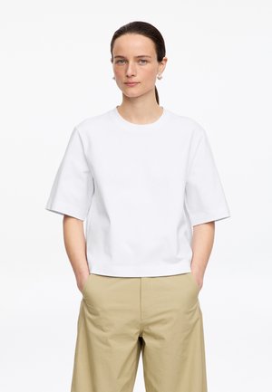 Jeune femme aux cheveux foncés attachés, portant un t-shirt blanc uni à manches courtes et un pantalon beige, les mains dans les poches, fond neutre.