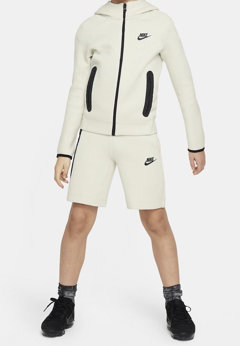 Sweat à capuche zippé blanc Nike avec accents noirs, associé à un short assorti. Comprend une capuche, des fermetures éclair et une finition texturée. Baskets noires portées.