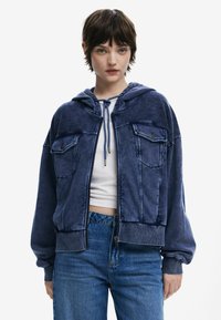 Blaues, kurz geschnittenes Hoodie aus weichem Stoff, mit zwei vorderen Taschen, Kapuze mit Kordelzug und gerippten Bündchen, kombiniert mit hochtaillierten Jeans.