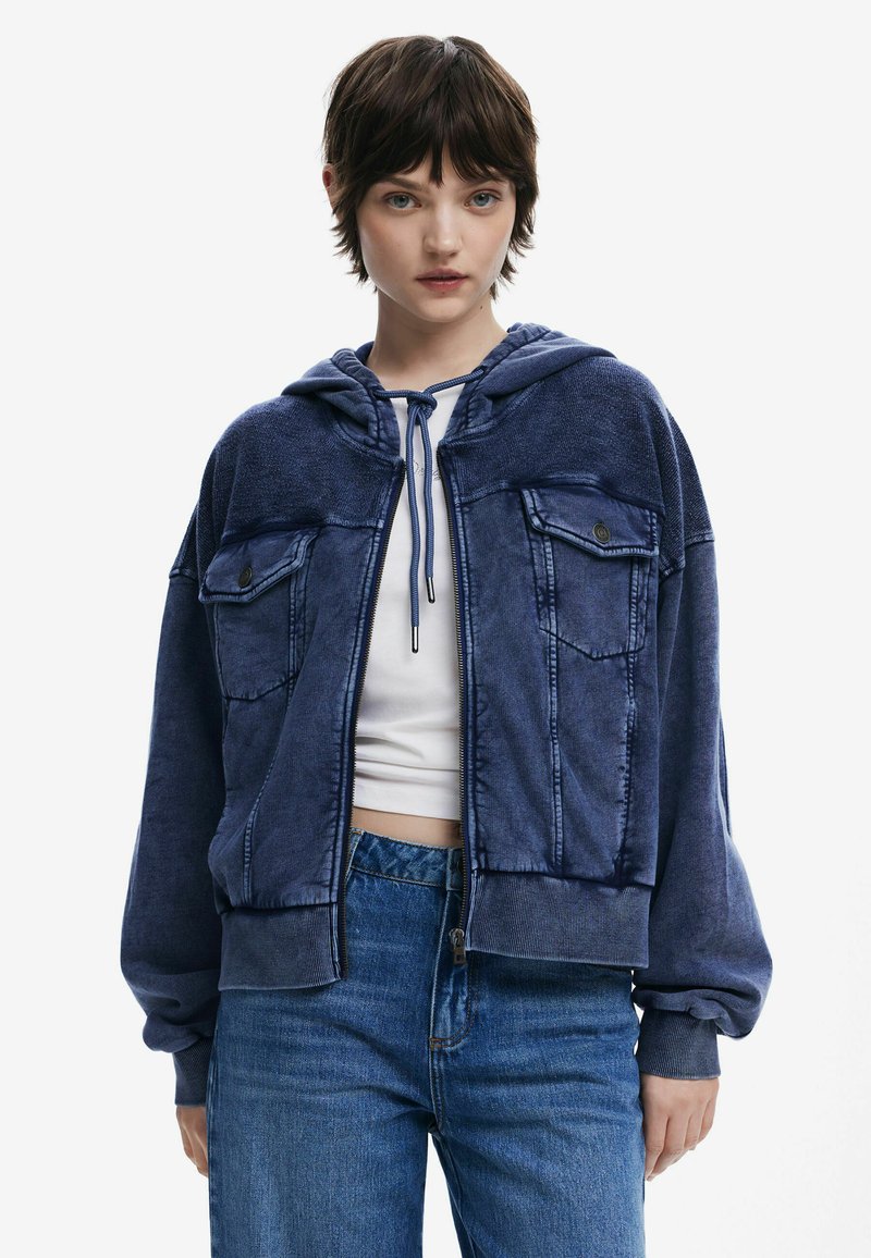 Blaues, kurz geschnittenes Hoodie aus weichem Stoff, mit zwei vorderen Taschen, Kapuze mit Kordelzug und gerippten Bündchen, kombiniert mit hochtaillierten Jeans.