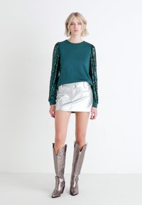 Sudadera verde azulado con mangas de lentejuelas, falda metálica plateada y botas vaqueras metálicas con diseños intrincados. Texturas suaves, forma ajustada.