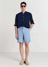 Marineblaue Hemdjacke aus strukturiertem Stoff, kombiniert mit hellblauen Shorts und beigen Slip-On-Schuhen, ergänzt durch dunkle Sonnenbrillen.