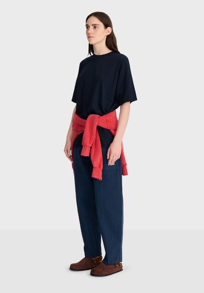 Jeune femme debout portant un t-shirt noir ample, un pantalon bleu marine, des chaussures marron à enfiler et un sweat-shirt rouge noué autour de la taille.