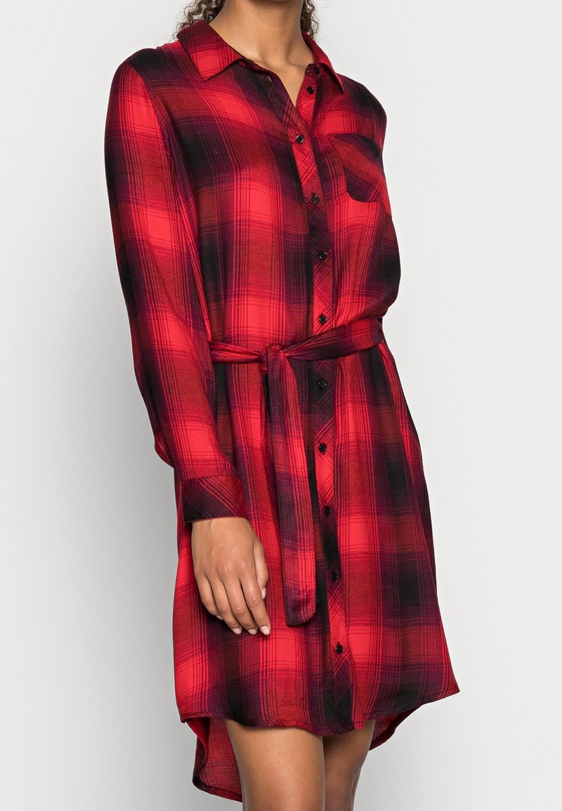 Femme portant une robe-chemise à manches longues à carreaux rouges et noirs, avec des boutons sur le devant et une ceinture à la taille, debout devant un fond uni.