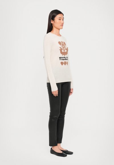 Ljusbeige stickad tröja med långa ärmar som har ett tryckt brunt och svart blommönster samt text. Kombinationen med svarta slim-fit byxor och ballerinas.