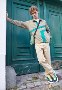 Chaqueta de forro polar marrón con acentos en teal, pantalones beige y zapatillas multicolores. Un hombre se apoya en una puerta verde, sosteniendo una pequeña bolsa verde.