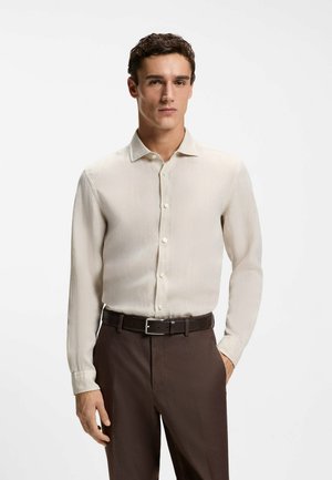 Mężczyzna o brązowych włosach ubrany w beżową koszulę z długim rękawem, wpuszczoną w brązowe spodnie z czarnym paskiem, stojący na białym tle.